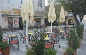 Schanigarten, © Konditorei Köller Ein leerer Schanigarten mit Tischen und Stühlen vor einem Café mit gelben Sonnenschirmen.