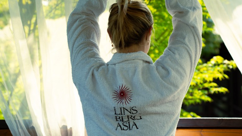 Yoga, © Asia Linsberg Person in einem weißen Bademantel mit dem Logo von Linsberg Asia, die Yoga vor einem Fenster mit Blick auf grüne Bäume praktiziert.