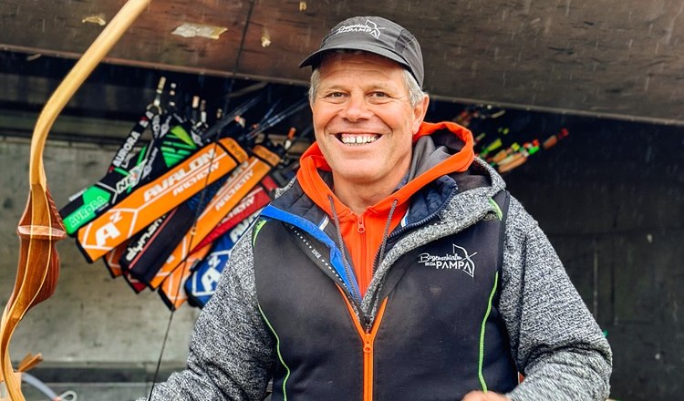 Bogenschiaßn in da Pampa, © Akademie am Trenkerhof Ein lächelnder Mann hält einen Bogen vor einem Anhänger mit Pfeilen.