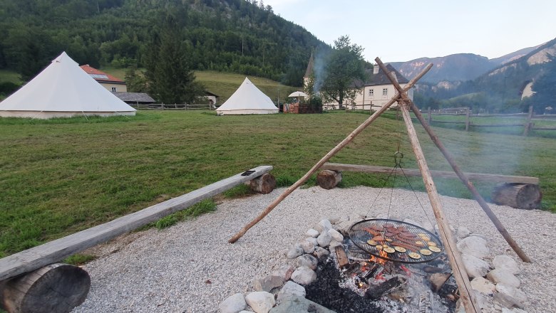 Feuerstelle - Wildnis Zentrum Nasswald, © Jonathan Clark Lagerfeuer mit Grill über Feuerstelle, Zelte im Hintergrund, umgeben von Bergen und Wiesen.