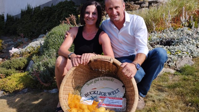 Martina and Richard Schwarz, © Fam. Schwarz A woman and a man sit smiling in a garden, holding a basket of potatoes and eggs.