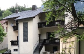 Ferienwohnung am Fluss, © Roland Krätzel Ein zweistöckiges Haus mit Balkon und Treppe, umgeben von einem grünen Garten mit Blumen.