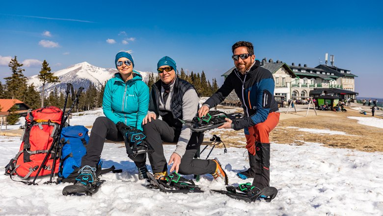 Geführte Schneeschuhwanderung, © Wiener Alpen/Kremsl Drei Personen sitzen im Schnee mit Schneeschuhen vor einem Berg und einem Gebäude.