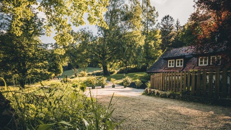 Landsitz Oberhof, © Niederösterreich Werbung / Maximilian Pawlikowsky Ein ländlicher Garten mit einem kleinen Haus und einem Kiesweg, umgeben von Bäumen und Pflanzen im Sonnenlicht.