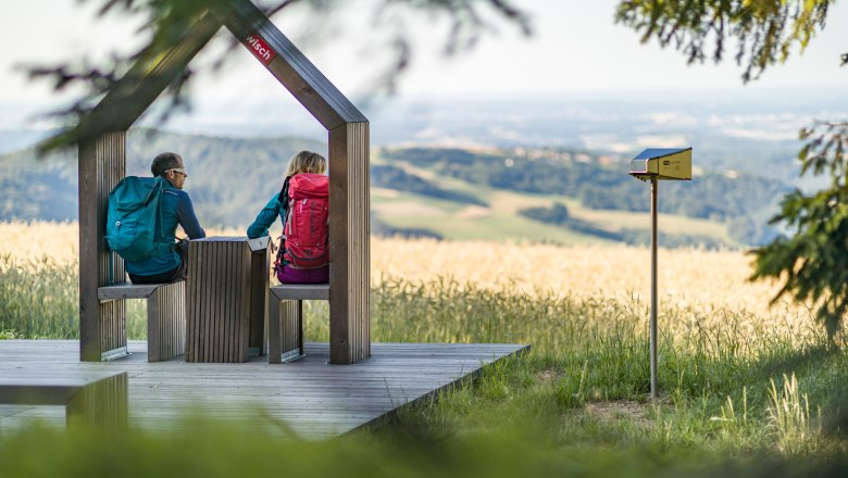Rastplatz in der Nähe der Aussichtswarte am Hutwisch, © Wiener Alpen/Martin Fülöp Zwei Wandernde sitzen auf einer Holzplattform mit Aussicht auf eine hügelige Landschaft.