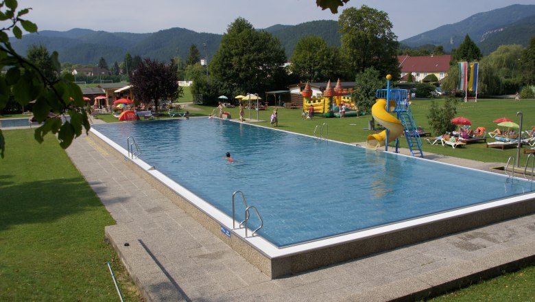 Freibad Waidmannsfeld, © Werner Zahornitzky Ein Freibad mit Schwimmbecken, Rutsche und Liegewiese, umgeben von Bäumen und Bergen.
