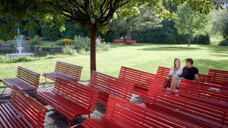 Musikpavillon Kurpark, © Wiener Alpen, Florian Lierzer Ein Paar sitzt auf roten Bänken im Kurpark, umgeben von Bäumen und einem Teich mit Springbrunnen.