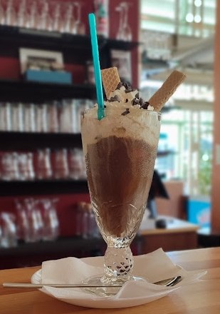 Cafe Elli, © Cafe Elli Ein Eiskaffee mit Schlagobers, Schokostreuseln und Waffeln in einem hohen Glas auf einem Tisch in einem Café.