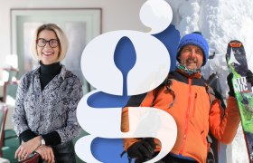 Hotel Schneeberghof mit Autor und "Hausmeister vom Schneeberg" Karl Tisch, © Wiener Alpen/Sophie Menegaldo, Karl Tisch Zwei Personen, eine Frau in einem Raum und ein Mann in Winterkleidung mit Skiern im Schnee, getrennt durch ein großes Logo.