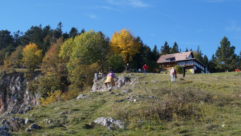 Wandern am Berg, © Reep Menschen wandern auf einem grasbewachsenen Hügel mit einer Berghütte im Hintergrund.