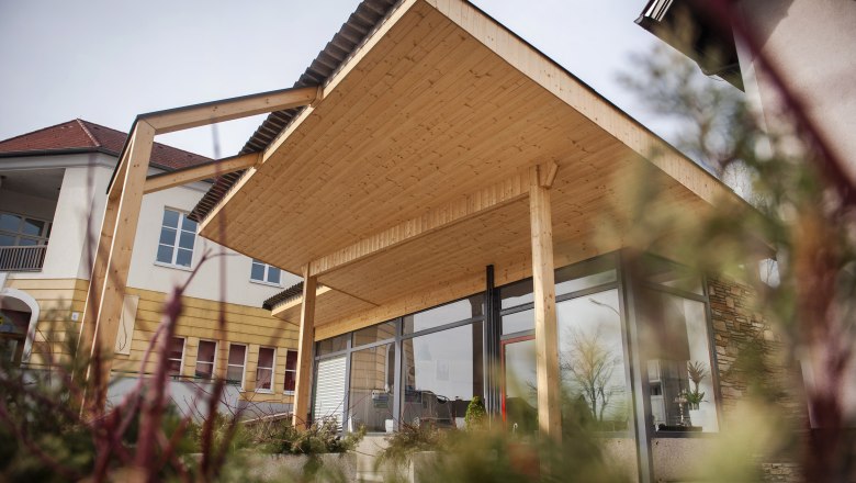 Info- & Genuss-Tankstelle Hochneukirchen, © Marktgemeinde Hochneukirchen / Stefan Knittel Modernes Gebäude mit Holzüberdachung und Glasfront, umgeben von Pflanzen.