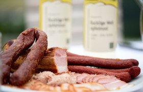 Mostheuriger Zierhof, © Wiener Alpen/Nicole Oberhofer Nahaufnahme von Wurst- und Fleischspezialitäten auf einem Teller mit Flaschen im Hintergrund.