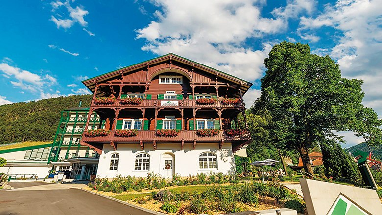 Schneeberghof in summer, © Hotel Schneeberhof A traditional, multi-storey building with wooden balconies and flowers, surrounded by trees and a blue sky.