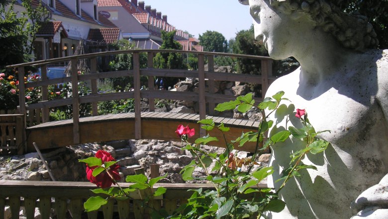Zum Griechen, © Trifinopoulos Dimtiros Steinstatue neben einer Holzbrücke und roten Rosen im Vordergrund.