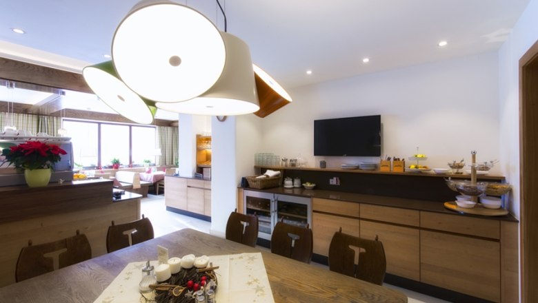 Das Restaurant, © Hotel Belvedere Ein modernes Restaurant mit Holzmöbeln, einem großen Tisch, dekorativen Lampen und einem Fernseher an der Wand.