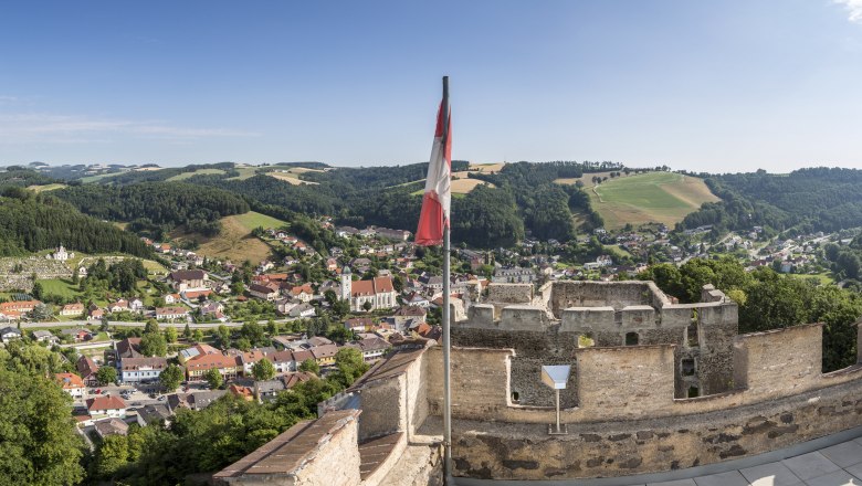 Blickplatz Feuerturm Burgruine, © Wiener Alpen, Foto: Franz Zwickl Panoramablick von einer Burgruine auf eine Stadt mit Hügeln im Hintergrund und einer österreichischen Flagge im Vordergrund.