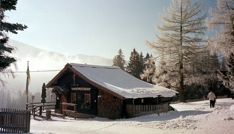 Grenzlandhütte, © Grenzlandhütte, Foto Paul Laschitz Eine verschneite Berghütte in winterlicher Landschaft mit Bäumen und einem Spaziergänger.