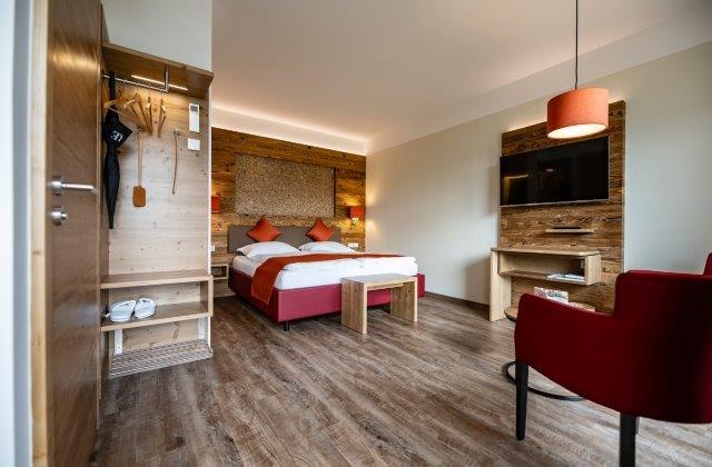 Das Gästehaus im Triad, © Marketing Platzhirsch, Gobauer Modernes Hotelzimmer mit Doppelbett, Holzmöbeln und rotem Sessel.