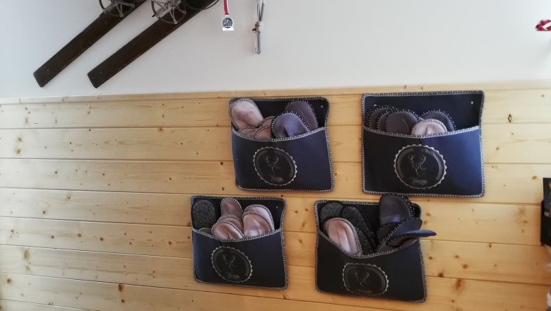 Eingang, © Familie Weibels Eingangsbereich mit Holzverkleidung, alten Skiern an der Wand und Filzpantoffeln in Taschen.