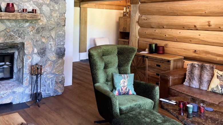 Wohnzimmer, © RaxChalet Gemütliches Wohnzimmer mit Steinwand, Kamin, grünem Sessel und Holzmöbeln.