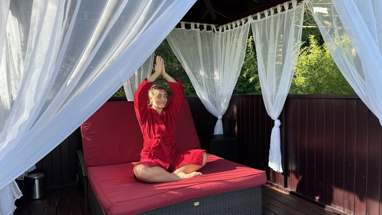 fernöstliche Gelassenheit, © Linsberg Asia Person in rotem Bademantel meditiert auf einer Liege in einem Pavillon mit weißen Vorhängen.