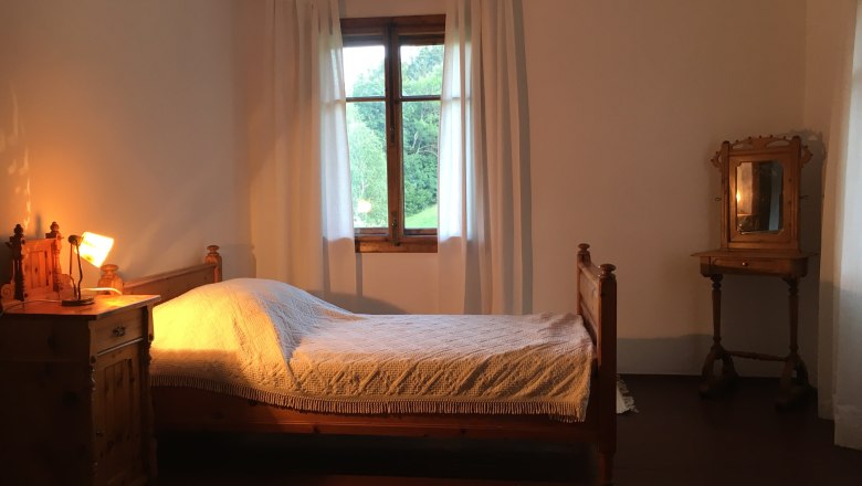 Ein rustikales Einzelzimmer mit Holzmöbeln, einem Bett, einem Nachttisch mit Lampe und einem Fenster mit weißen Vorhängen.