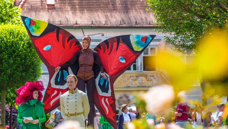 Bunt verkleidete Menschen beim Rosenfest, eine Frau ist auf Stelzen als großer bunter Schmetterling verkleidet