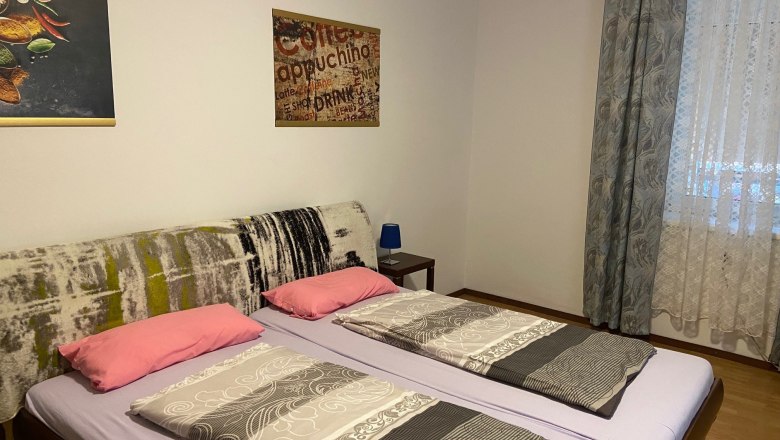 Schlafzimmer mit Doppelbett, zwei Kissen, zwei Decken, Wandbilder und Fenster mit Vorhängen.