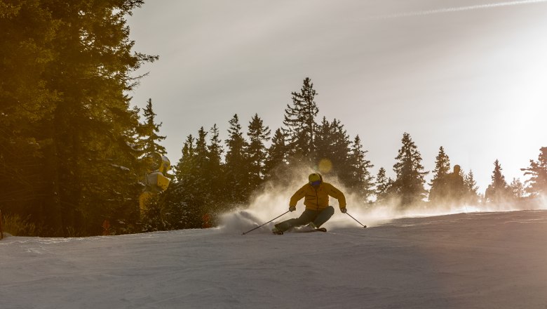 Skifahrer auf einer Piste bei Sonnenuntergang, umgeben von Bäumen.
