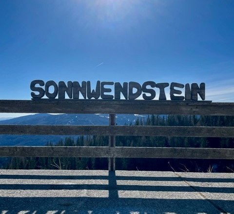 Großbuchstaben mit 'Sonnwendstein' vor sonnigem Himmel und Berglandschaft.