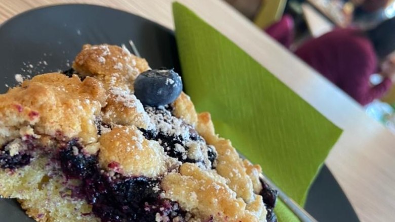 A slice of blueberry cake on a black plate with fresh blueberries and powdered sugar, with a blurred café scene in the background.