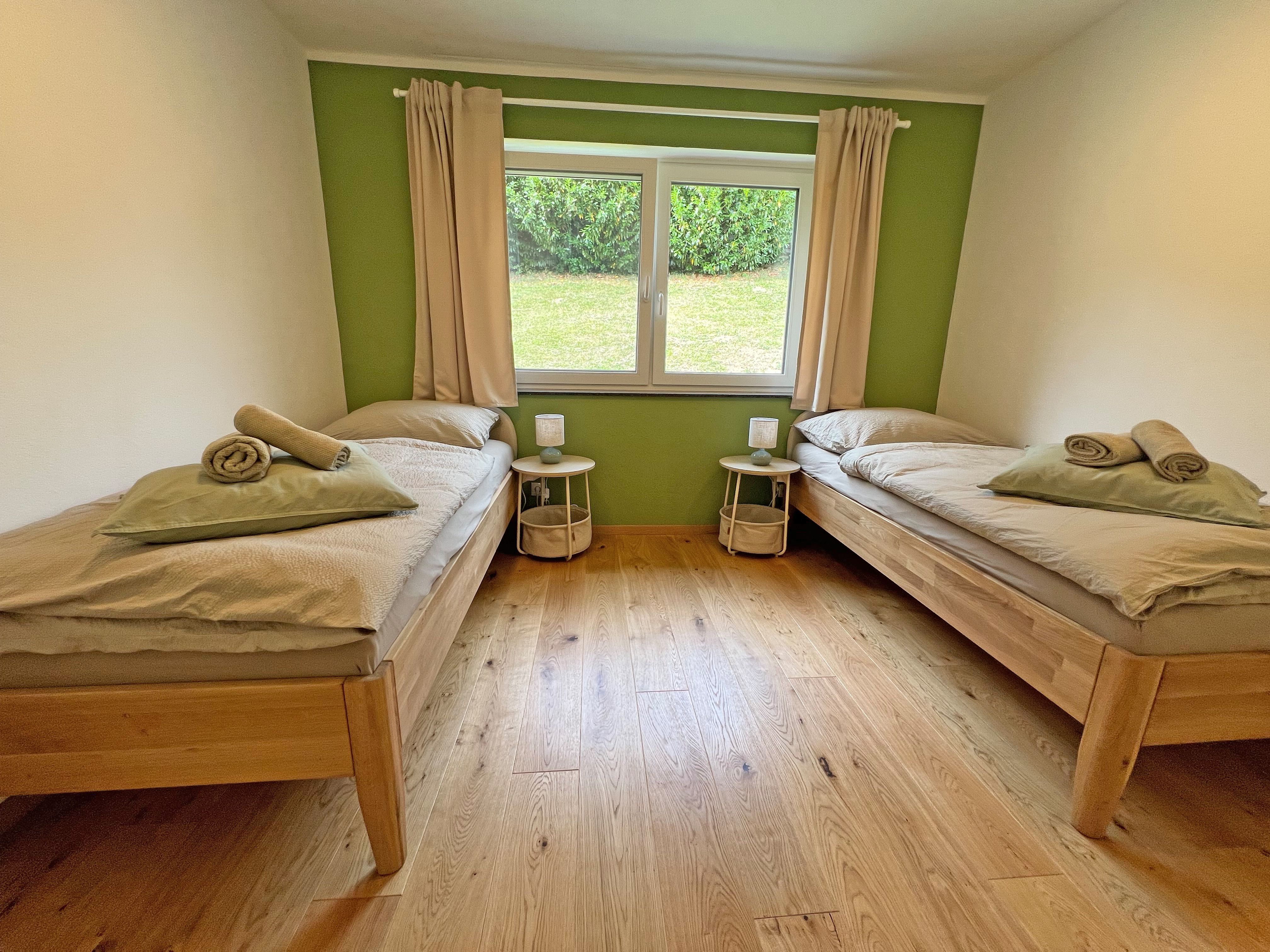IMG_3692: A bright bedroom with two single beds, a window wall painted green, hardwood floors, and a view of the garden.