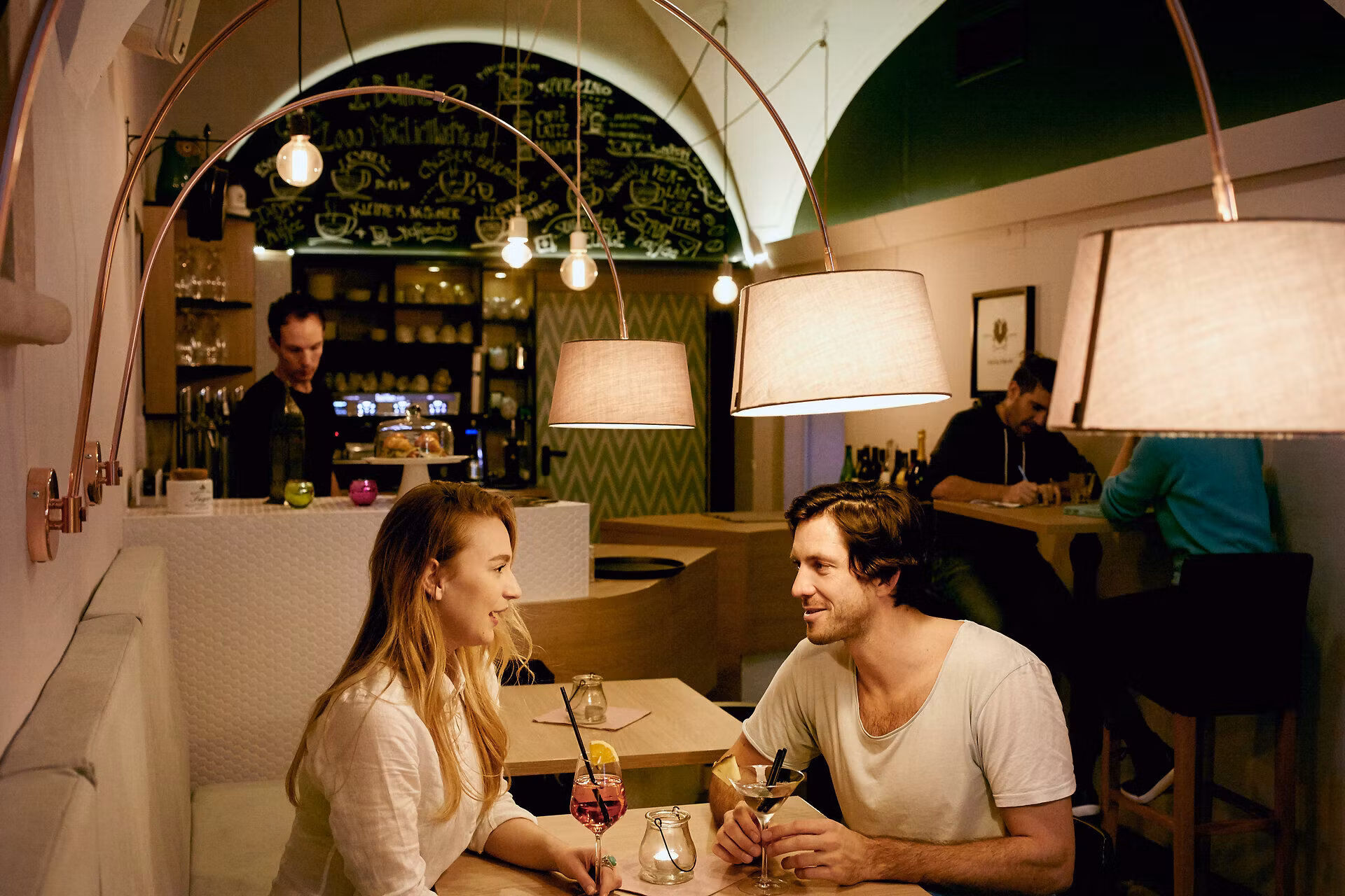 Ein Pärchen sitzt auf einem Tisch in einem Lokal mit Weinglas und Martiniglas und stoßen an.