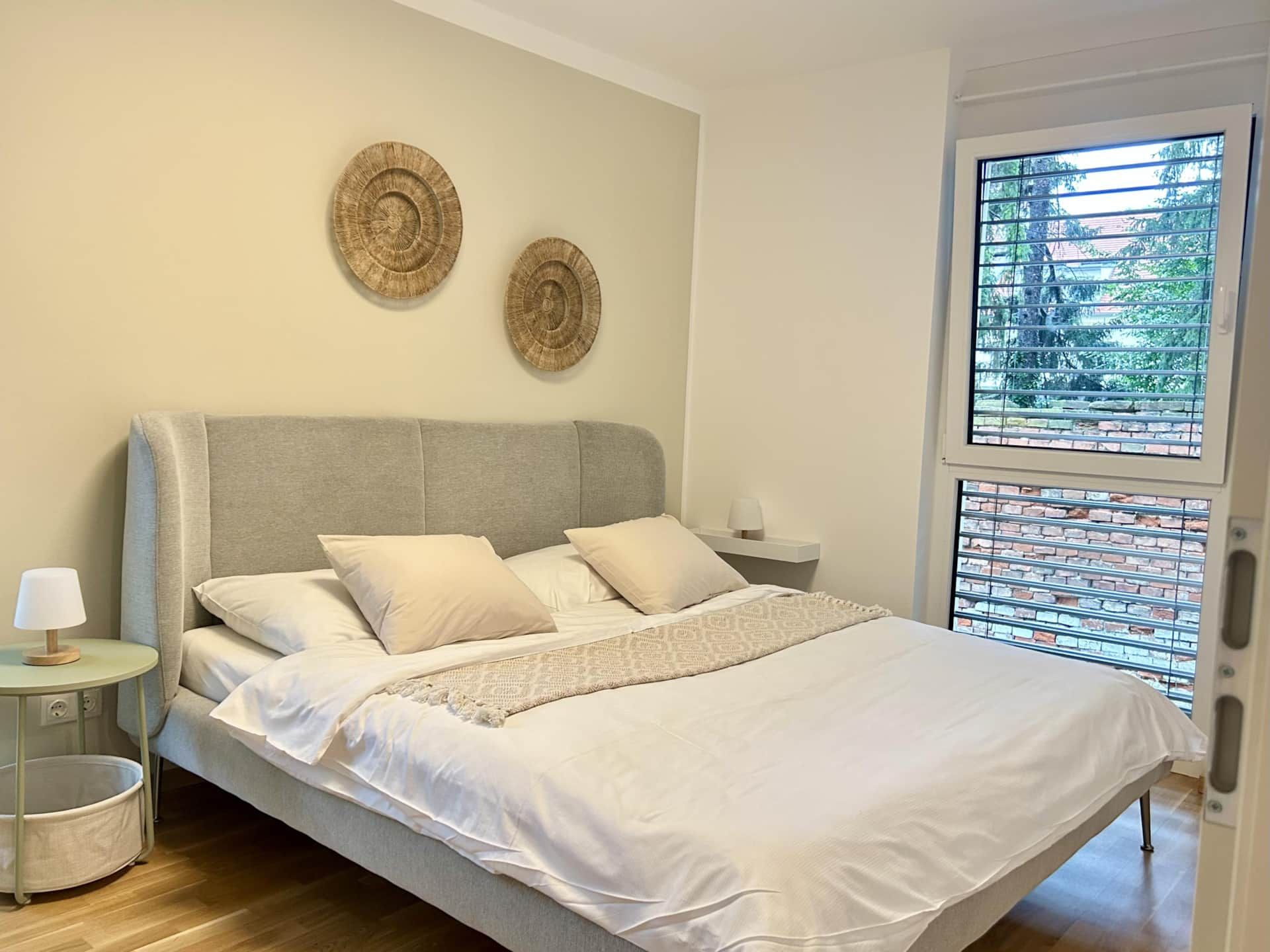Cozy bedroom with fabric bed, bedside table, large window and boho-style wall decoration