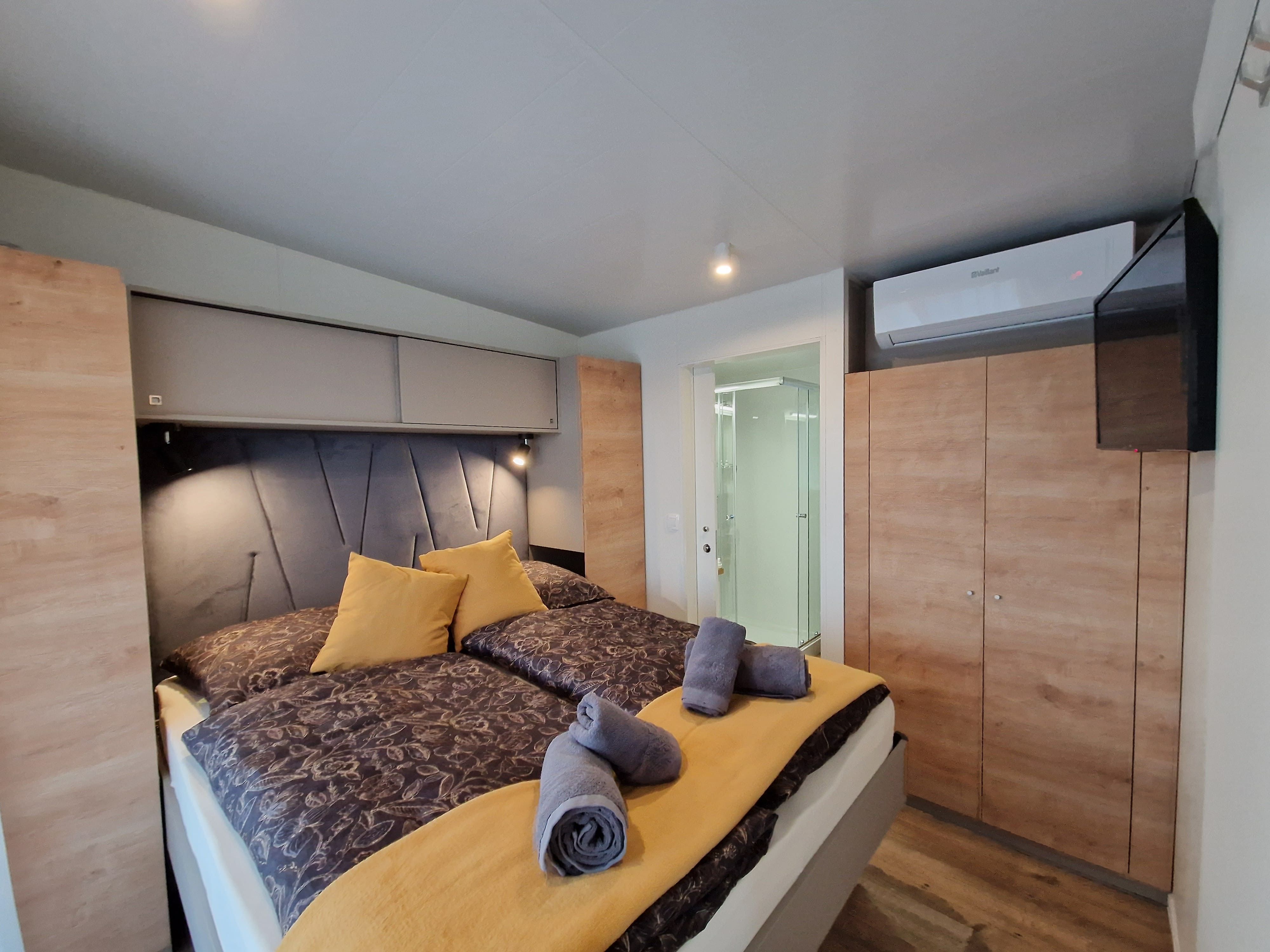 Innenansicht eines Schlafzimmers mit Doppelbett, gelben Kissen und Handtüchern, Schrank, Klimaanlage und Blick ins Badezimmer.