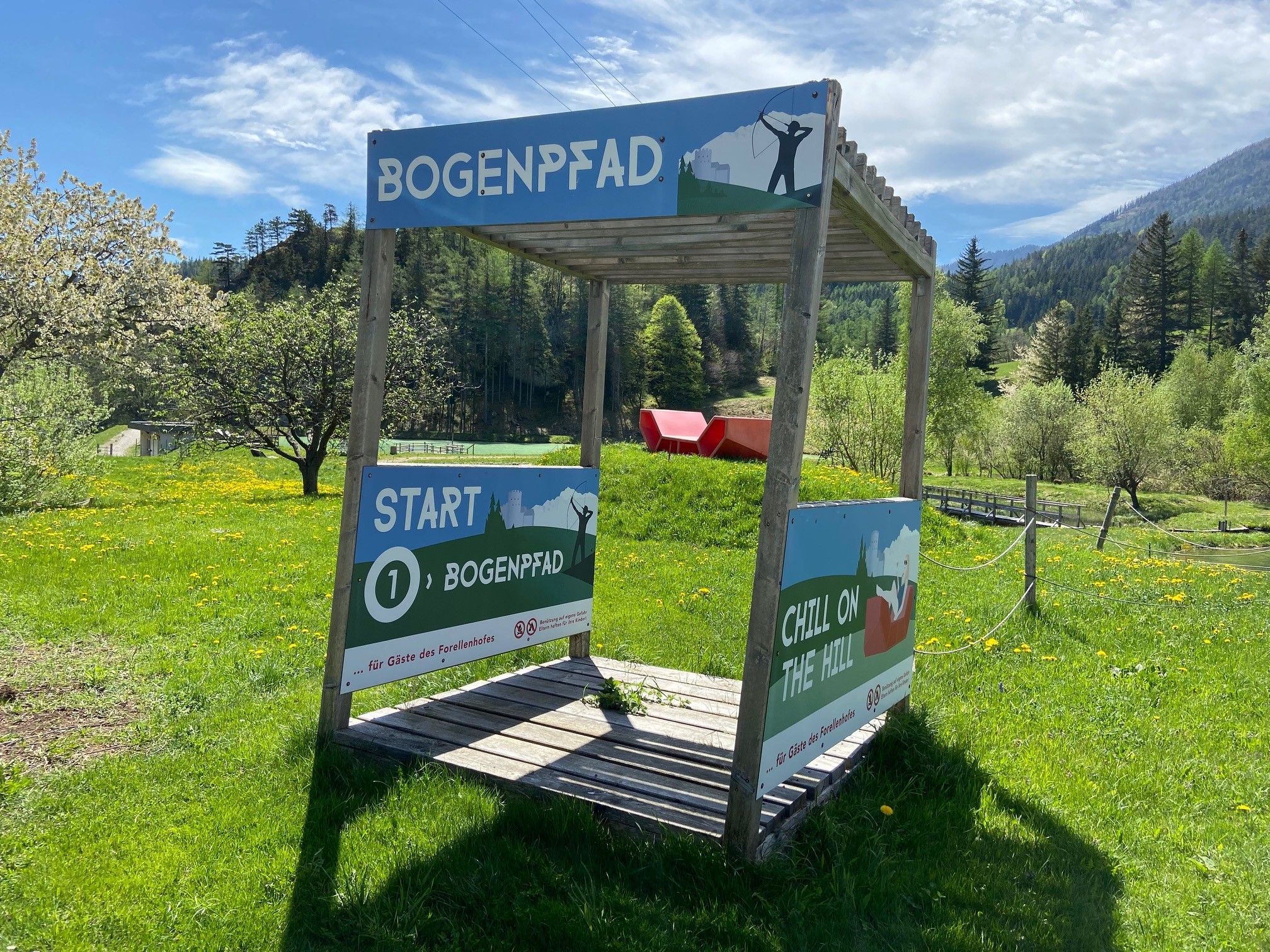 Startpunkt des Bogenpfads mit Schildern auf einer grünen Wiese, umgeben von Bäumen und Bergen.