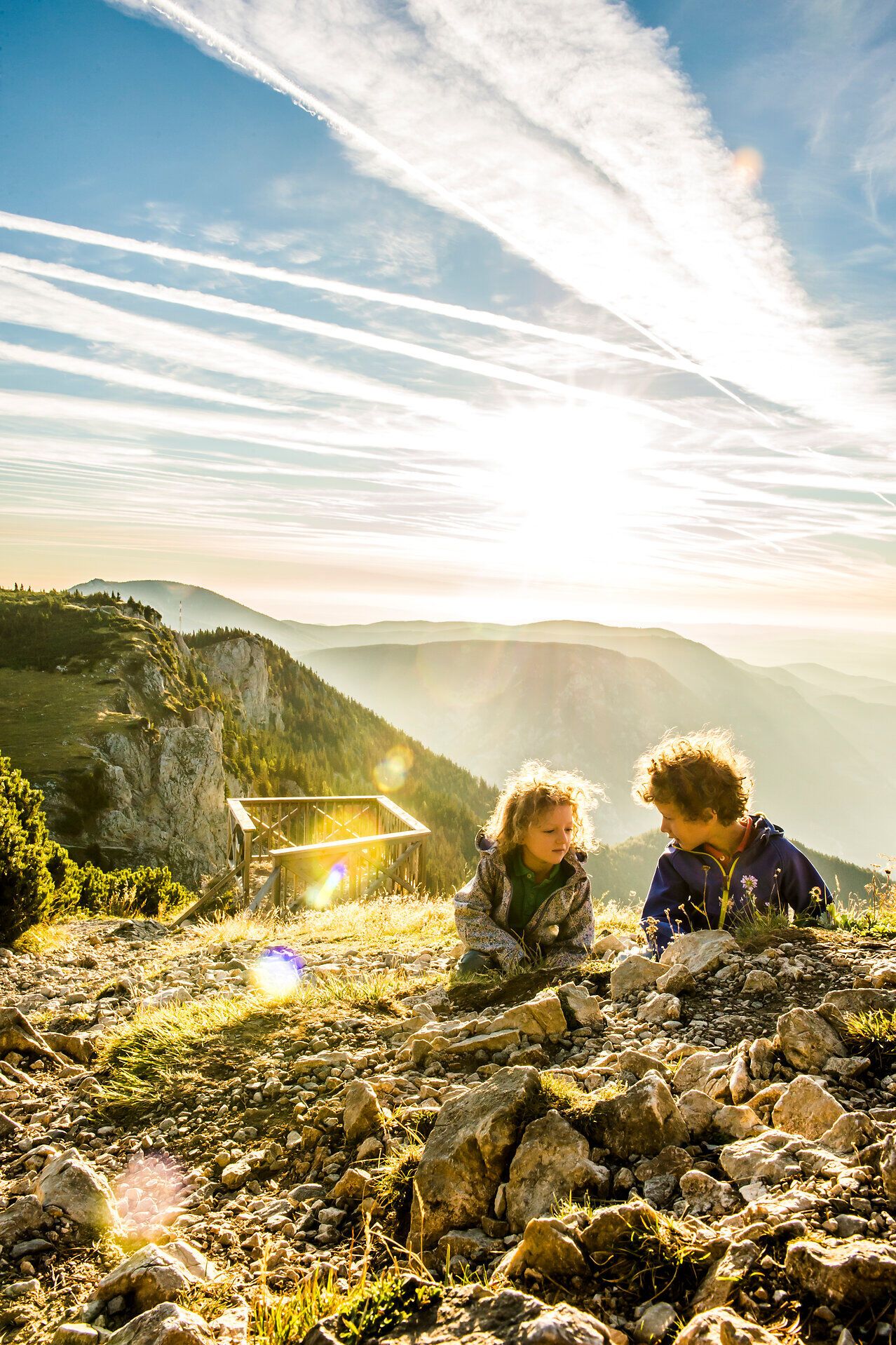 In der warmen Abendsonne sitzen zwei Kinder auf dem sanften, steinigen Hang und genießen die atemberaubende Aussicht auf die umliegenden Berge. Die goldenen Strahlen des Sonnenuntergangs tauchen die Landschaft in ein magisches Licht und schaffen eine unvergessliche Atmosphäre der Freude und des Staunens.
