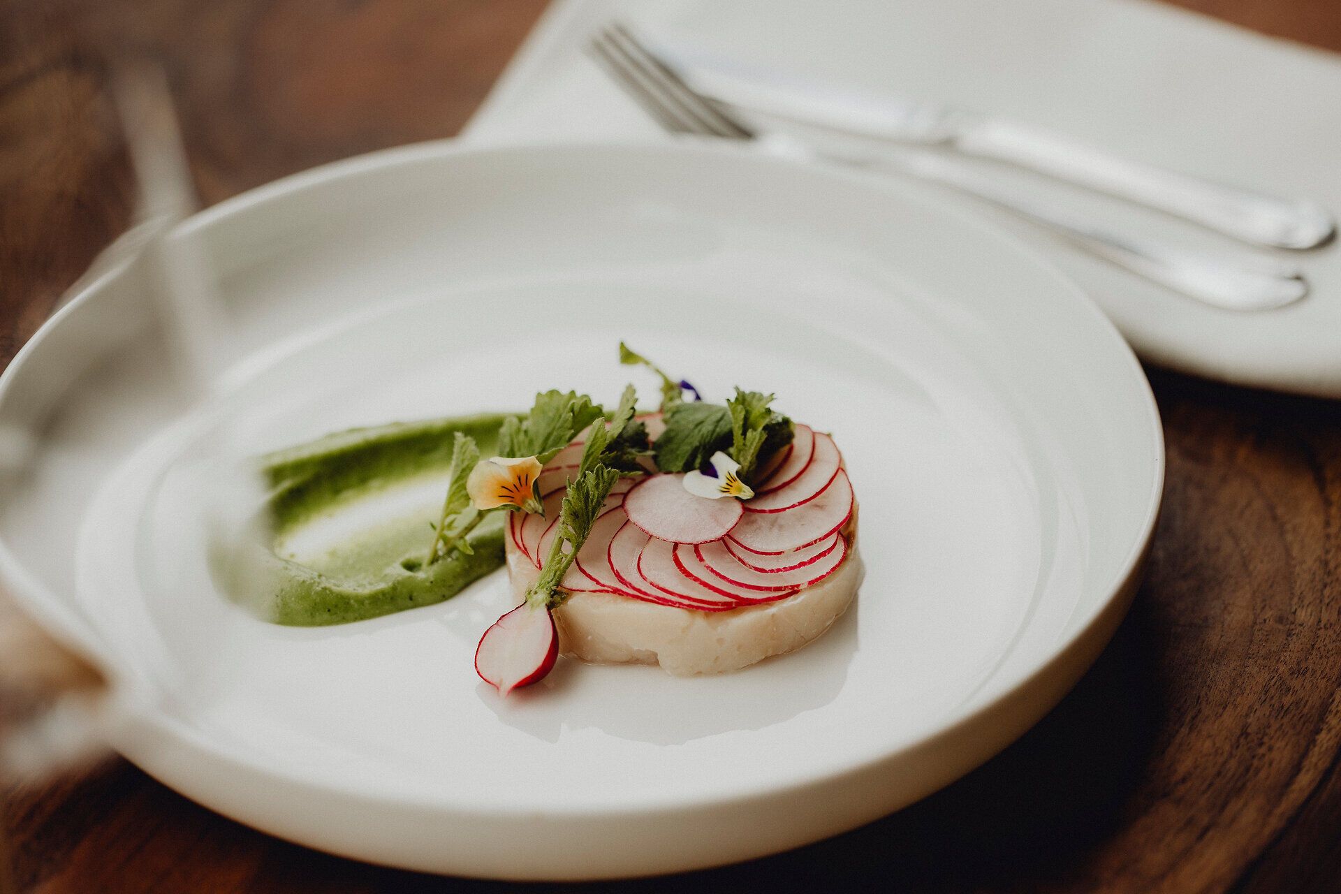 Ein kunstvoll angerichteter Teller präsentiert eine delikate Kombination aus zartem Saibling, frischen Radieschen und einer cremigen Wasabi-Sauce. Die leuchtenden Farben der Zutaten und die filigranen Kräuter verleihen dem Gericht eine einladende Frische, die zum Genießen einlädt.