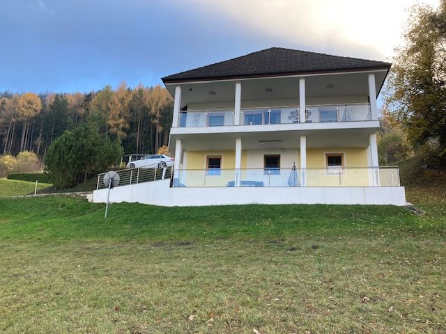 Zweistöckiges Haus mit Balkon vor einem Wald.