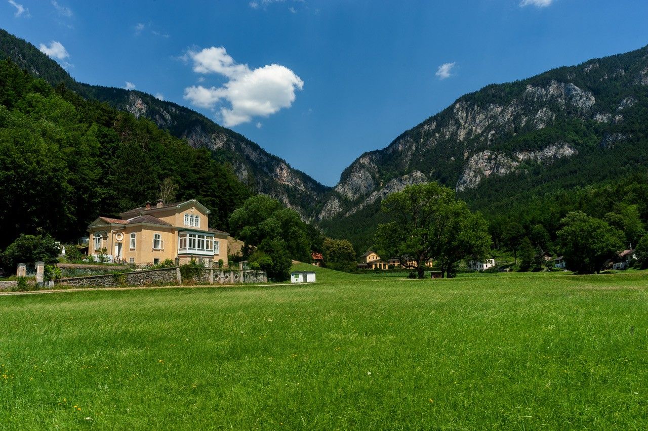 Ein gelbes Haus vor einer Bergkulisse mit grüner Wiese im Vordergrund.