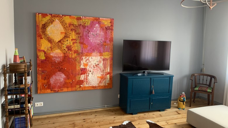 Wohnzimmer mit gro&szlig;em buntem Gem&auml;lde, Fernseher auf blauem Schrank, Holzboden und Kuhfell-Teppich.