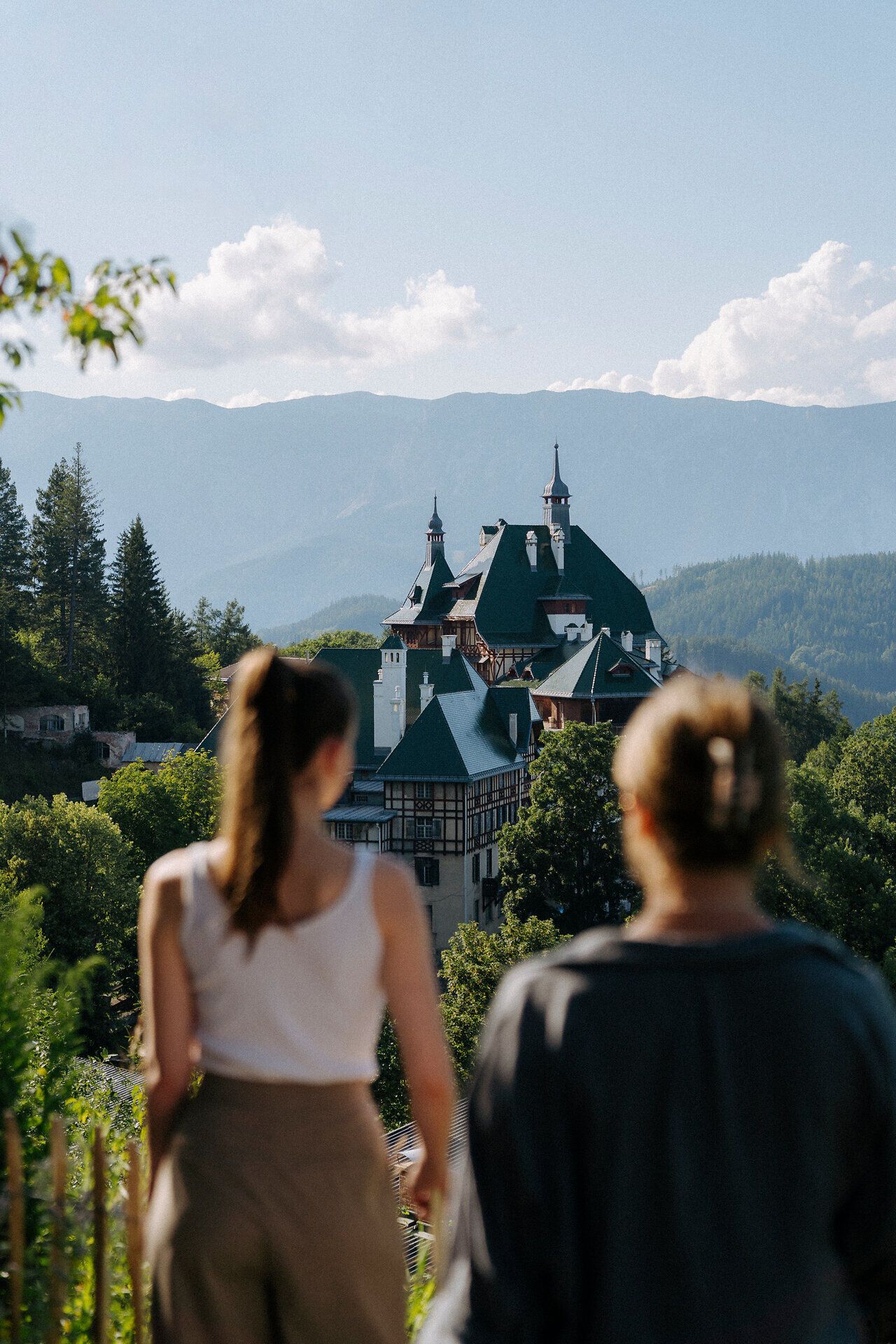 Zwei Menschen, mit dem Rücken zur Kamera, blicken auf eine malerische Berglandschaft. Im Vordergrund sieht man das Südbahnhotel, eingebettet in üppiges Grün. Im Hintergrund erheben sich Berge unter einem klaren blauen Himmel.