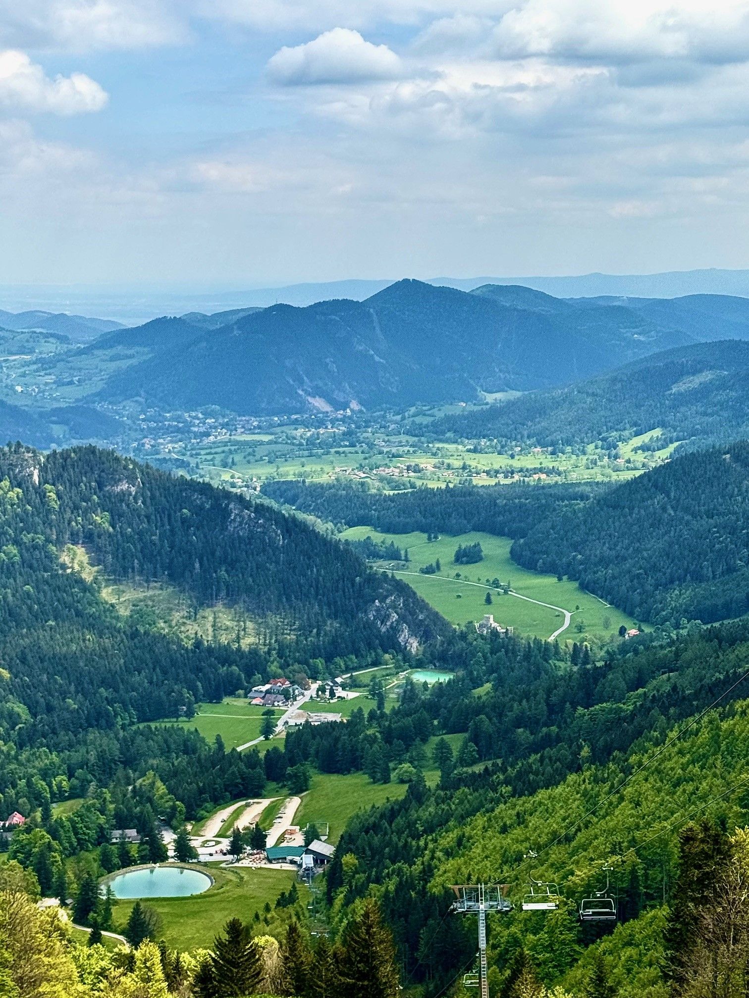 Landschaft mit grünen Hügeln, Wäldern und einem kleinen Dorf im Tal, umgeben von Bergen unter einem bewölkten Himmel.