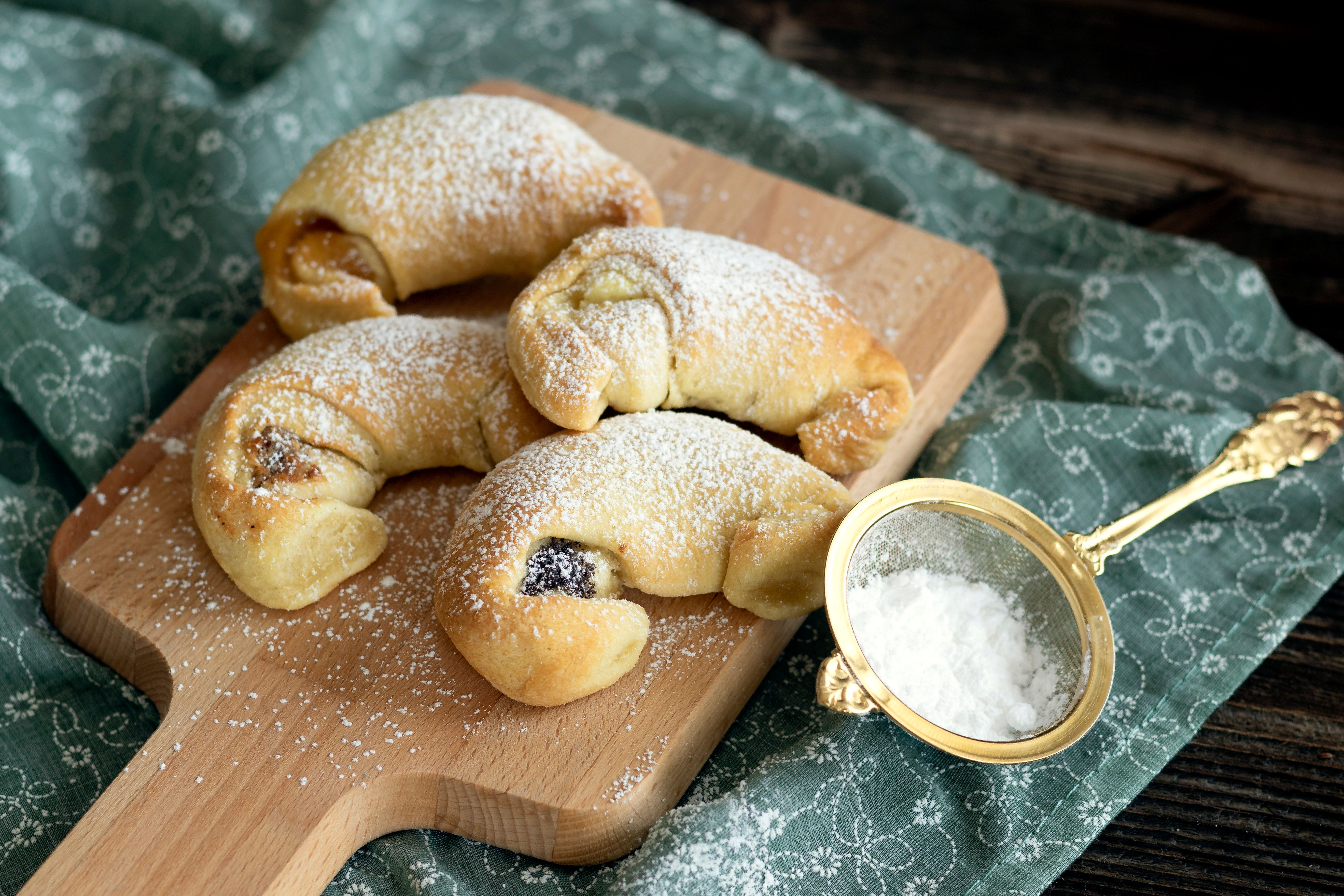 Vier Rahmkipferl auf einem Holzbrett mit Puderzucker bestreut, daneben ein Sieb mit Puderzucker.