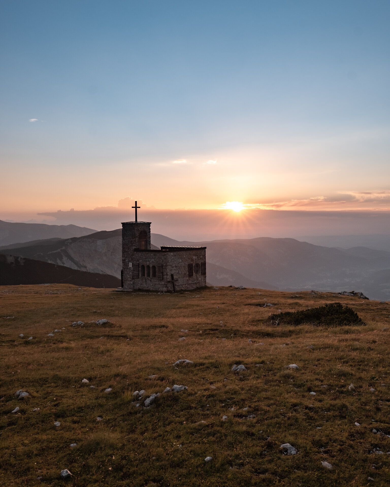 Die sanften Hügel der Wiener Alpen umarmen die alte Kapelle, während die Sonne langsam hinter den Bergen verschwindet. Ein goldener Schimmer breitet sich über die Landschaft aus und lädt Wanderer ein, die Ruhe und Schönheit der Natur zu genießen.