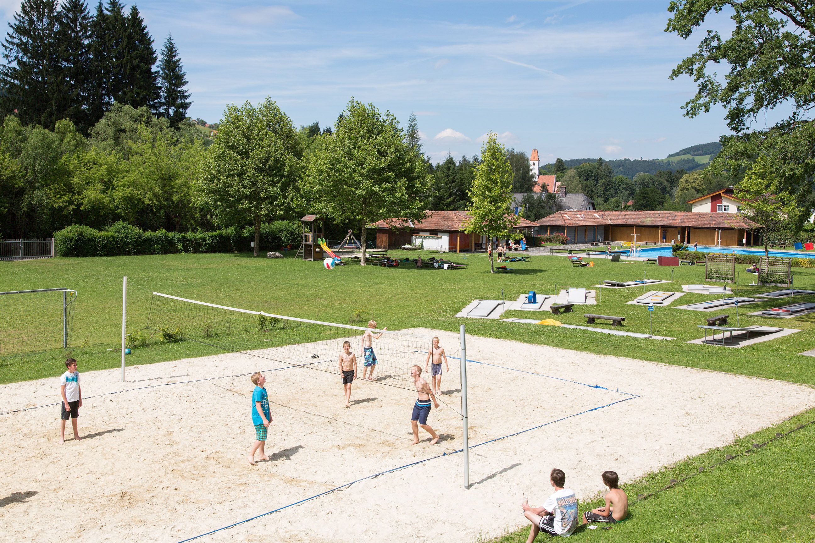 Kinder spielen Volleyball auf einem Sandplatz im Freibad Aspang, umgeben von grüner Wiese und Bäumen.