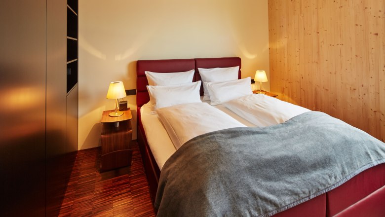 Útulná ložnice s manželskou postelí, červeným čelem, dvěma nočními stolky s lampičkami a dřevěnými stěnami.