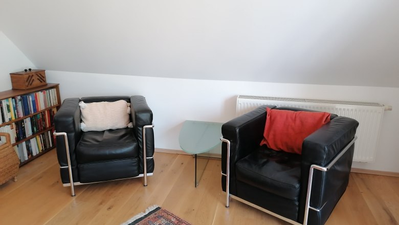 Zwei schwarze Ledersessel mit Kissen, ein Glas-Beistelltisch, B&uuml;cherregal und Teppich in einem Wohnbereich mit Dachschr&auml;ge.