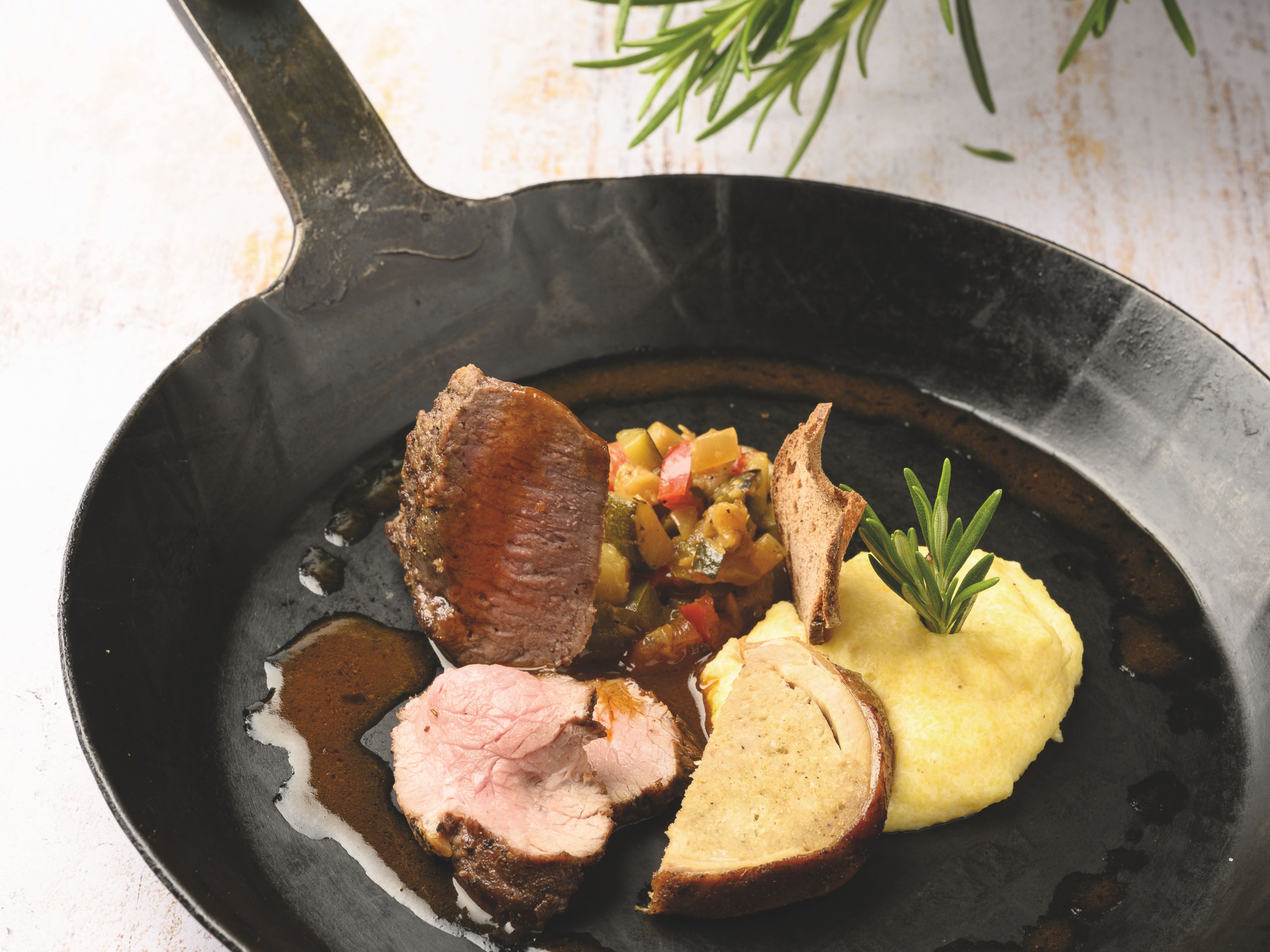 Ein Gericht mit verschiedenen gebratenen Fleischstücken, Kartoffelpüre und Gemüse in einer Pfanne serviert.