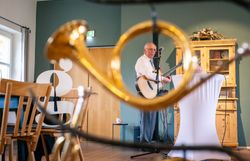 Musiker mit Gitarre durch ein Blasinstrument fotografiert steht in einer Gaststube mit Blick in die Kamera